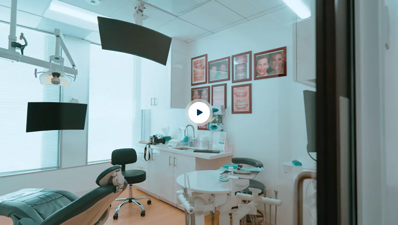 Modern dental examination room with reclining patient chair, dentist stool, overhead light, two black monitors, white cabinets, sink, and framed photos of smiles on the wall.