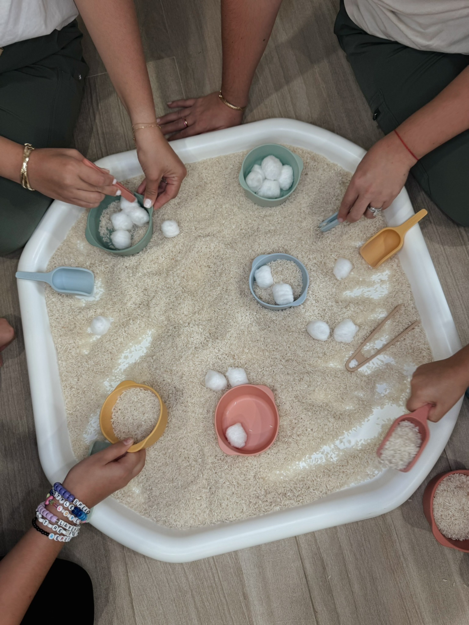 Hands explore a rice-filled sensory bin with scoops, bowls, and cotton balls.