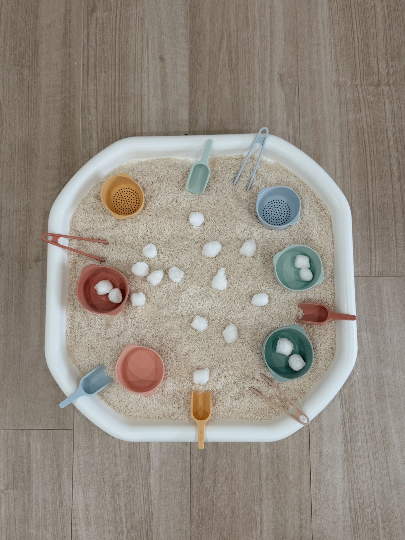 A sensory bin prepared with rice, cotton balls, bowls, scoops, and tongs.