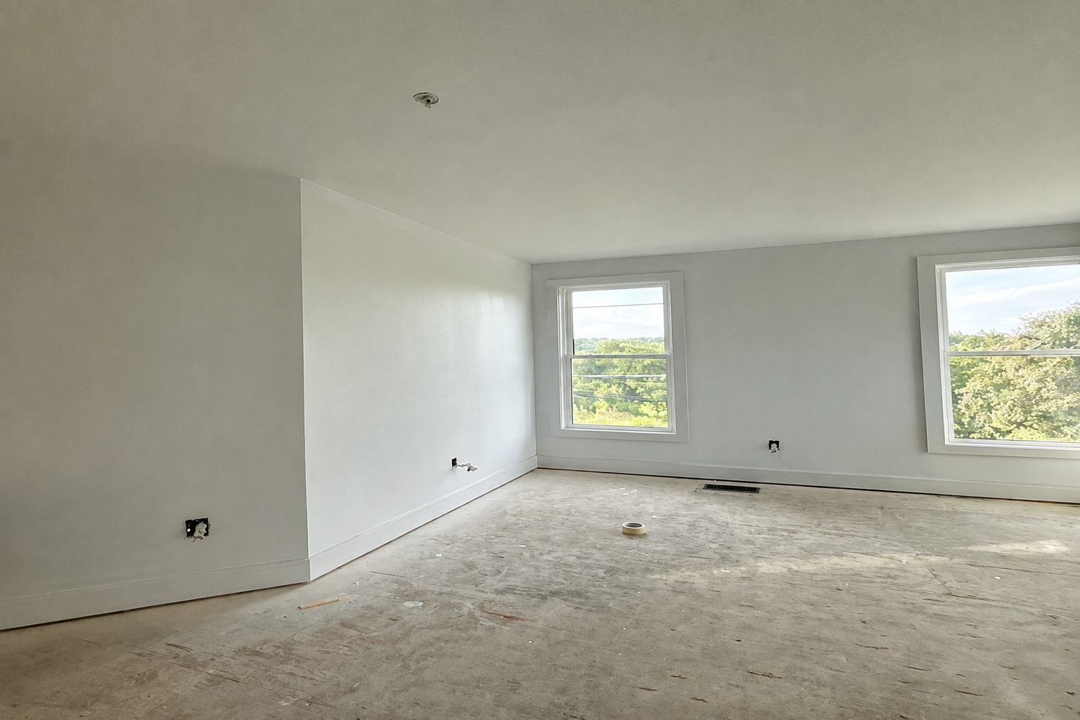 Empty room with freshly painted light-gray walls, white trim, two windows, and an unfinished floor.
