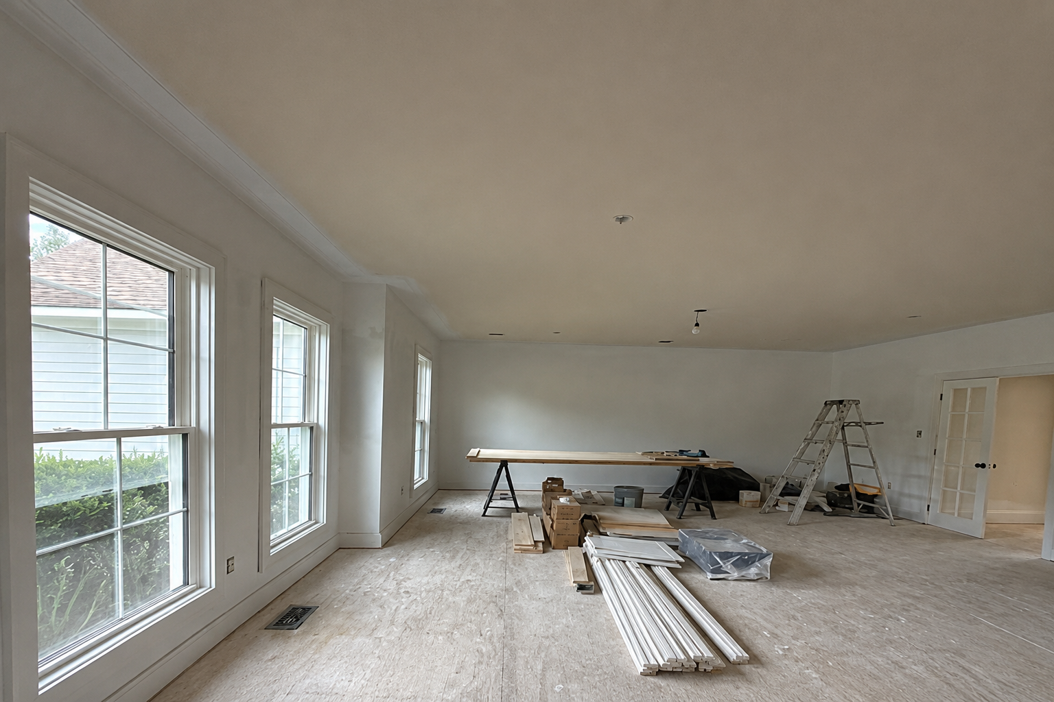 Interior painting project in progress with a ladder, worktable, tools, and stacked trim inside a large room.