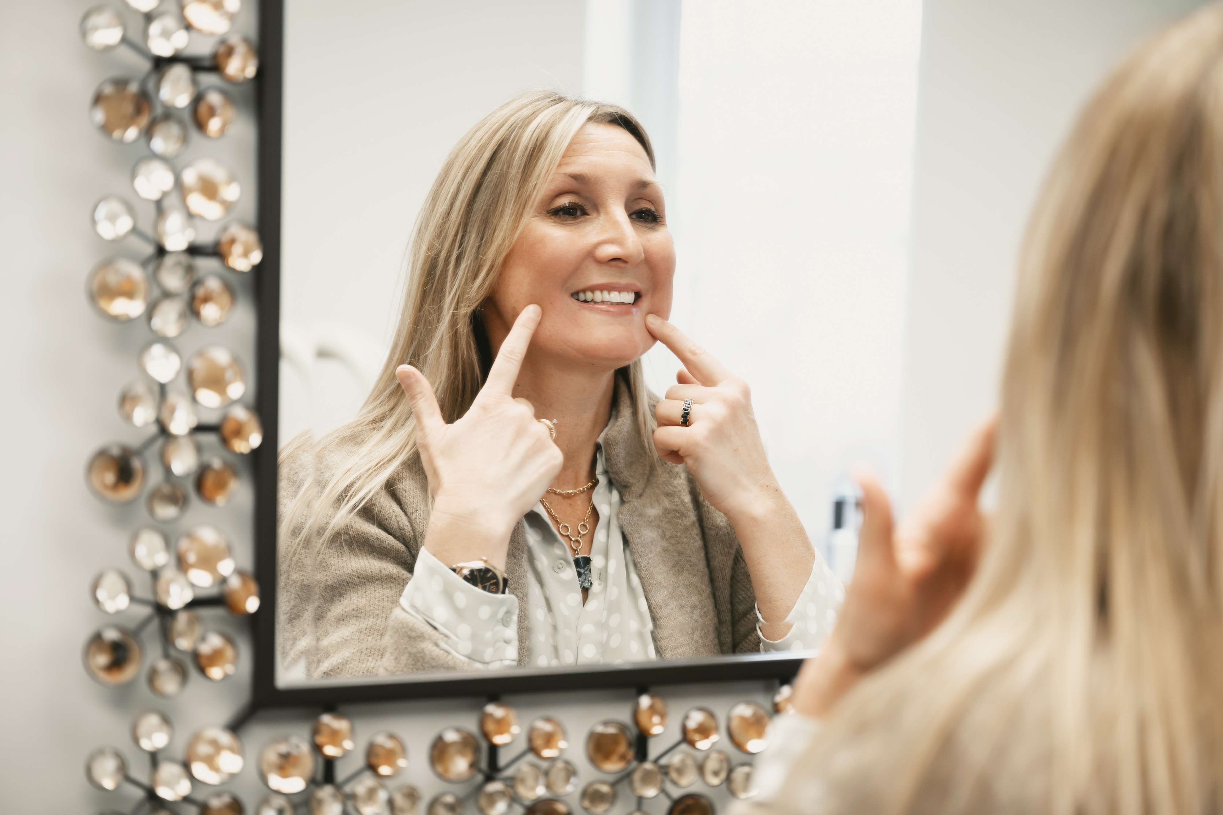 A blonde woman smiling in the mirror and pointing to her smiles