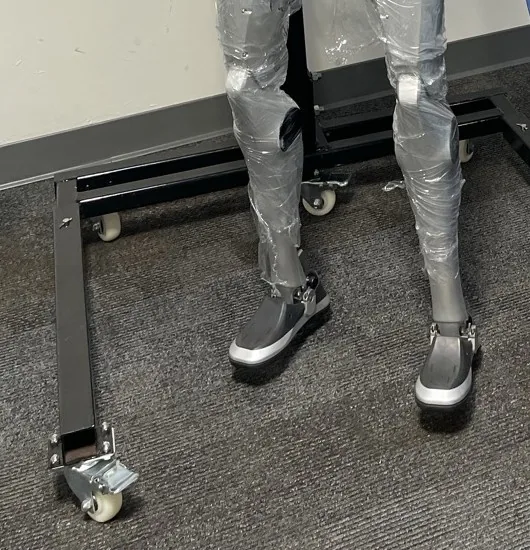 Close view of the plastic-wrapped legs and feet of a humanoid robot standing on a castered steel support frame on carpet.
