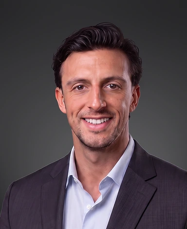 Smiling man with dark hair wearing a dark blazer and light blue dress shirt against a dark gray background.