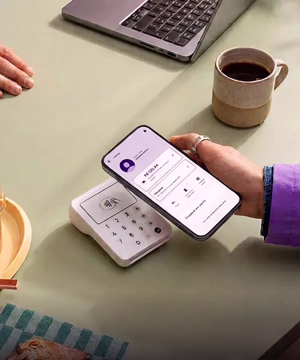 Pessoa segurando celular para pagamento sem contato perto de máquina, com laptop e xícara de café na mesa.