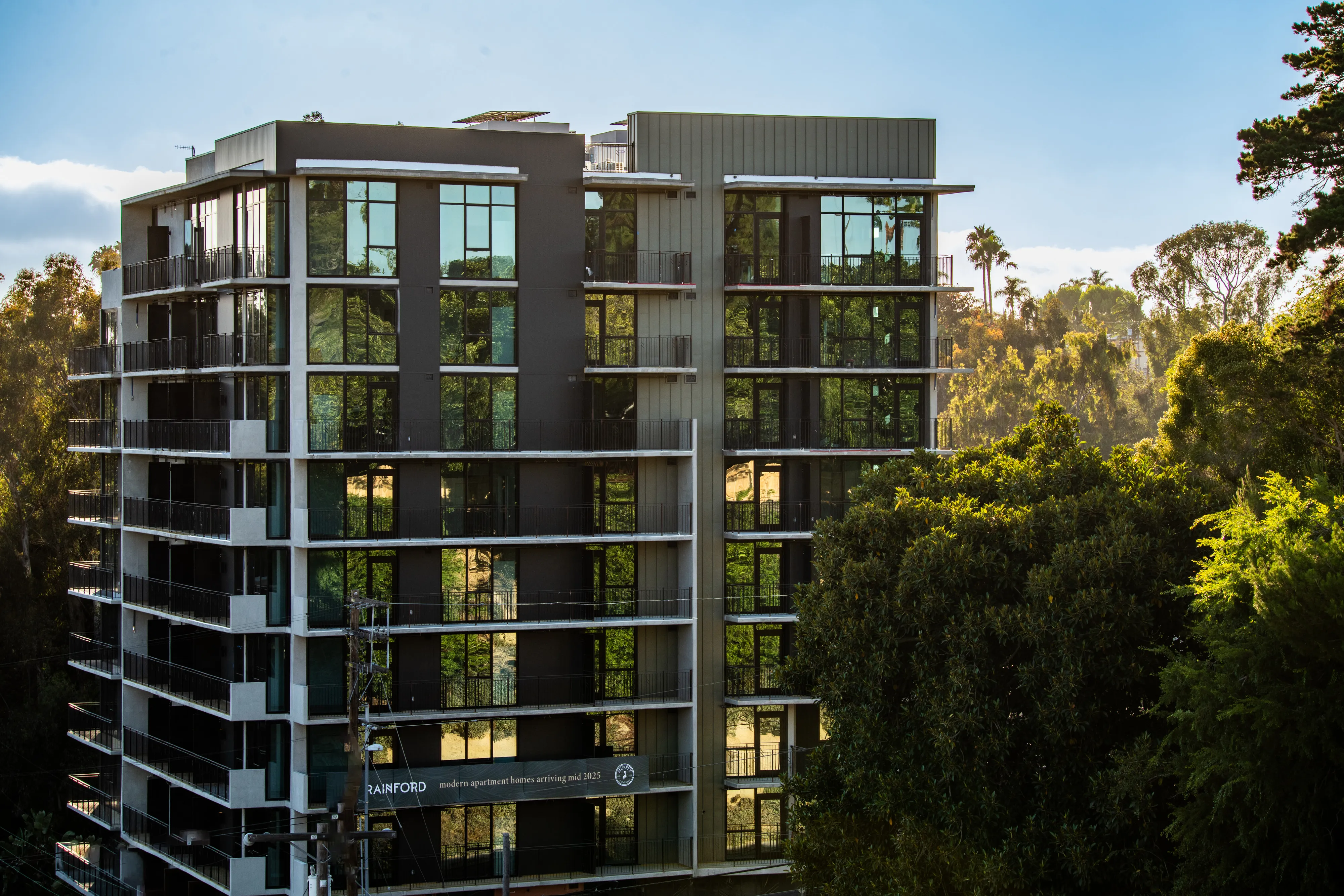 Modern multi-story apartment building surrounded by lush green trees under a clear blue sky.