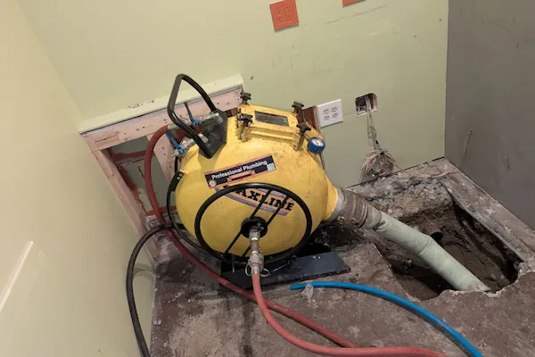 Yellow professional plumbing machine connected to red and black hoses beside a floor opening with exposed pipe in a room under repair.