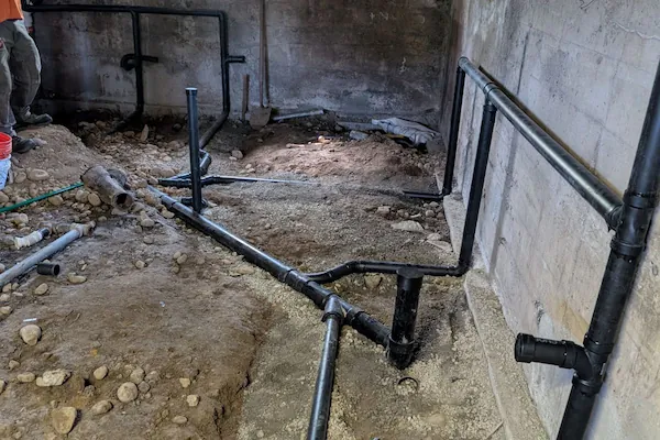 Basement under construction with exposed black plumbing pipes installed along the dirt floor and concrete walls.