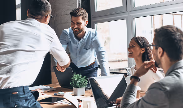 Two men shaking hands across a meeting table while colleagues smile in the background.