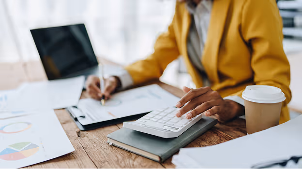 Person in a yellow blazer using a calculator while reviewing charts and taking notes at a desk.
