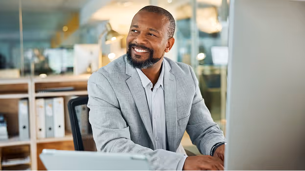 Smiling man in a grey suit sitting at his desk in a modern office.