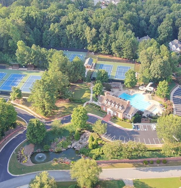 Aerial view of a community pool, tennis courts, clubhouse, parking, and trees in a neighborhood.