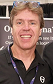 Smiling man with blond hair wearing a black zipped jacket in front of a dark background.