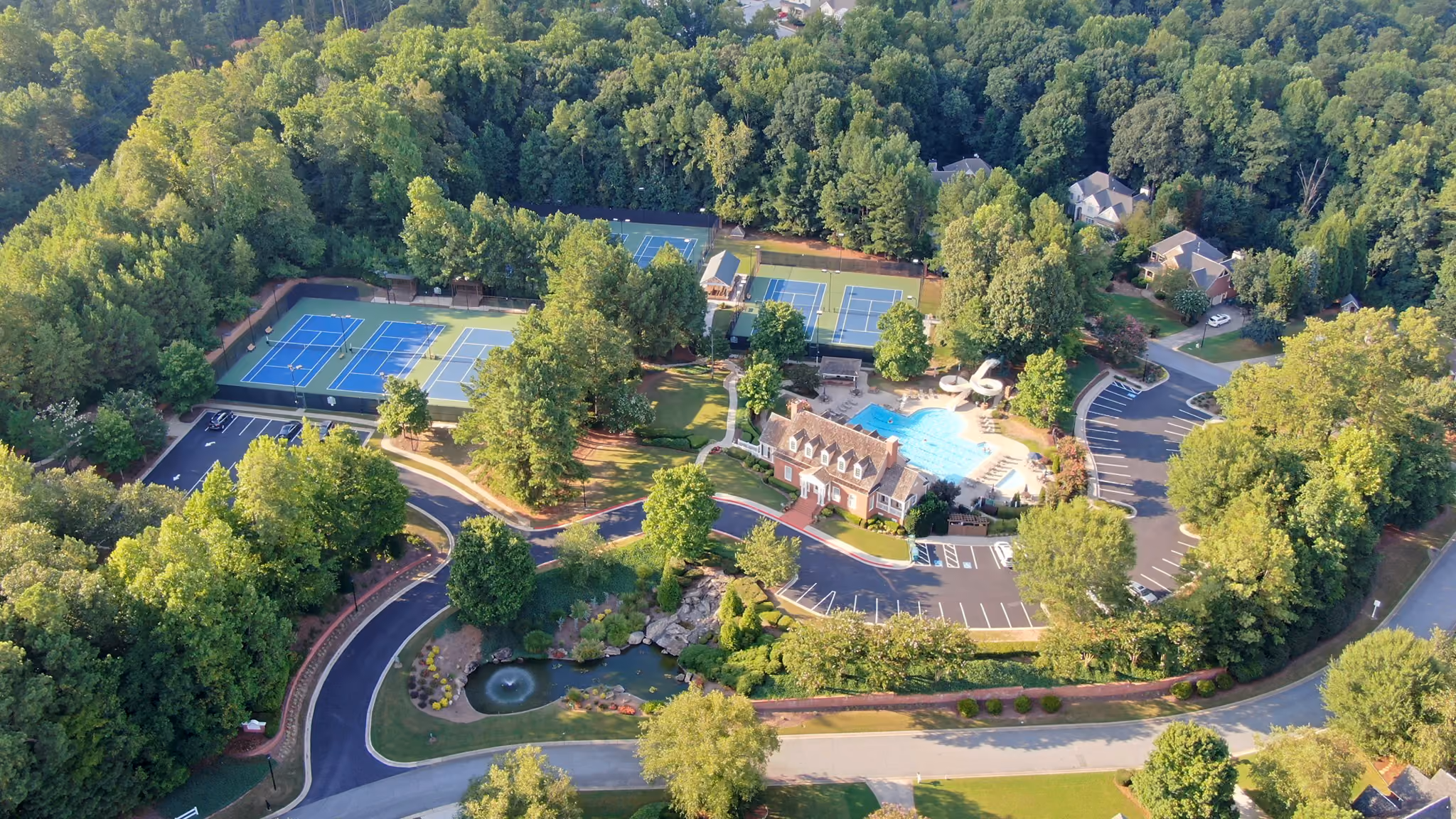 Aerial view of a forested neighborhood area featuring a community recreation center with a large swimming pool, multiple tennis courts, a small pond with a fountain, and surrounding parking lots and roads.