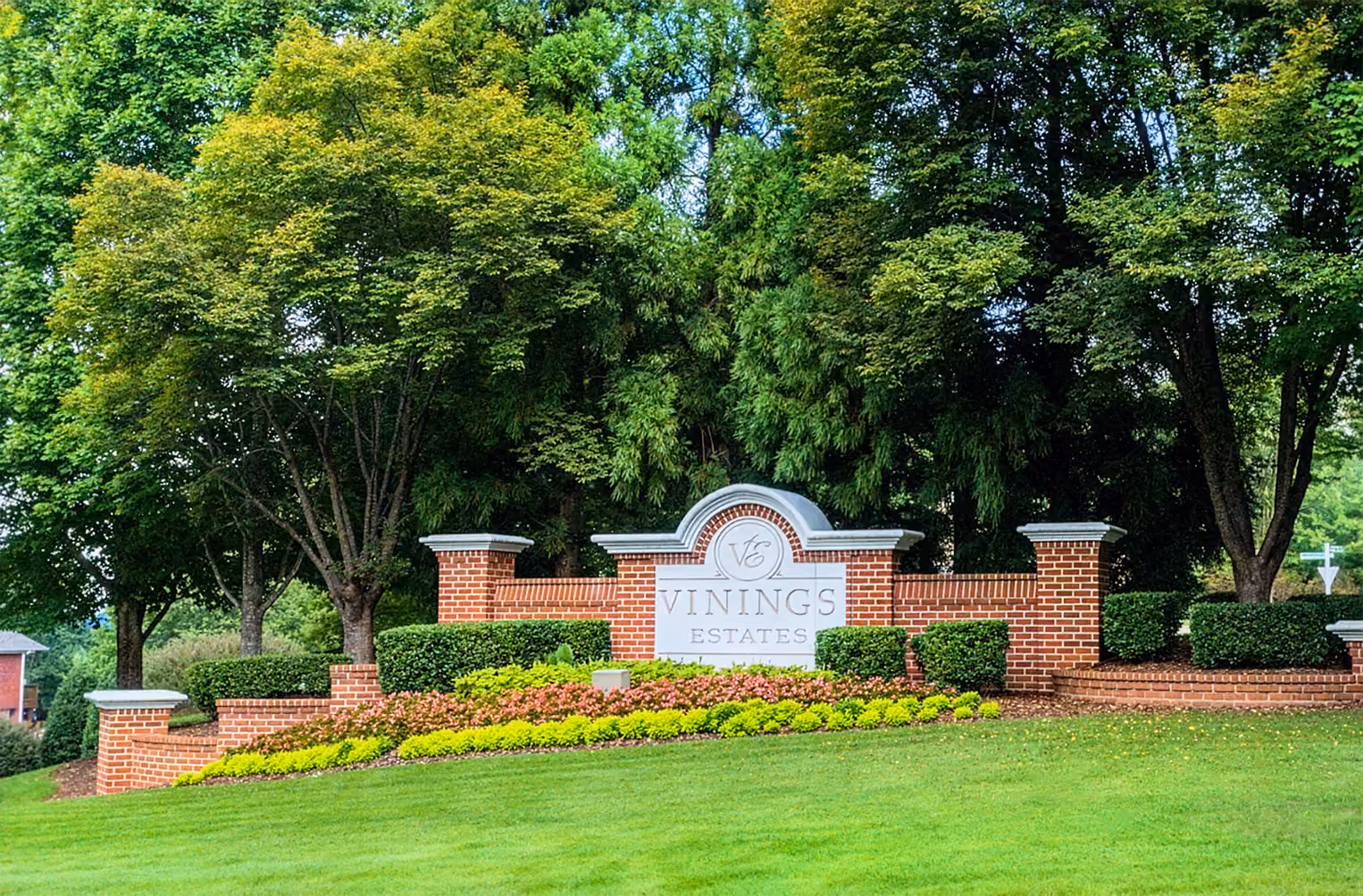 Brick entrance sign with white center panel reading "Vinings Estates" surrounded by trimmed bushes, flowering plants, and tall green trees behind it.