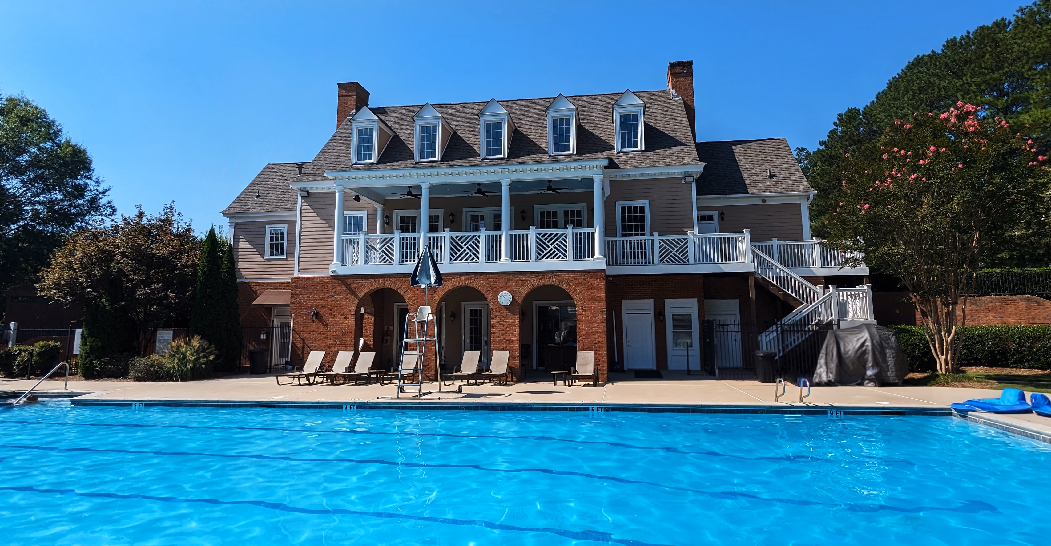 Outdoor swimming pool with clear blue water in front of a large two-story clubhouse featuring a brick lower level, beige upper level with white trim, multiple dormer windows, and a covered balcony with white railings.