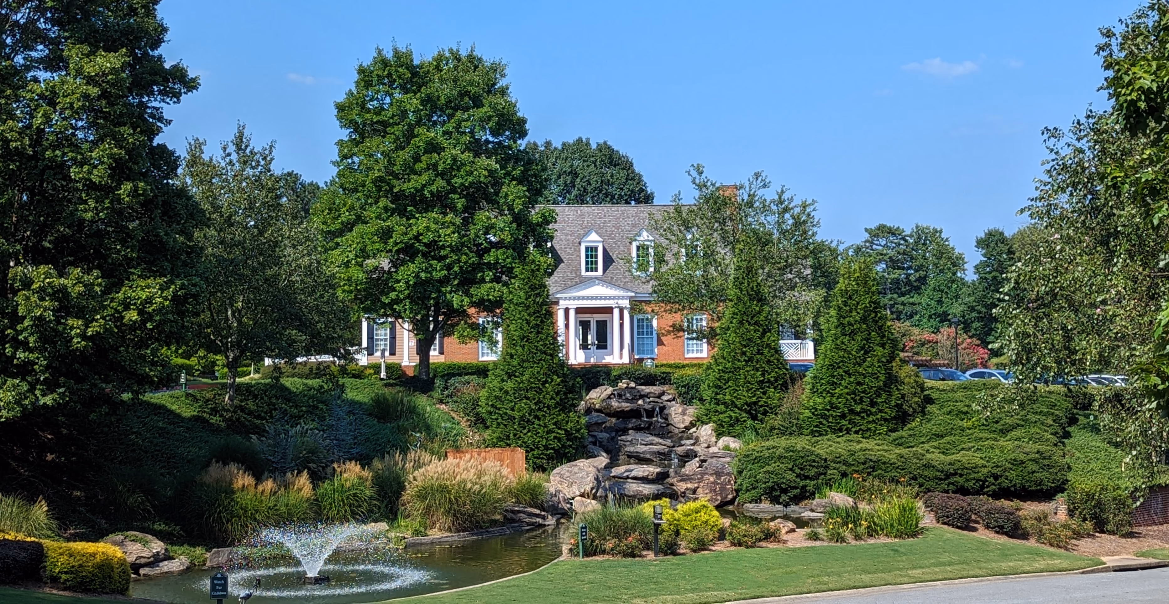 Large brick house with white columns and dormer windows surrounded by lush green trees, shrubs, and a manicured lawn, featuring a rocky waterfall and a pond with a water fountain in front.