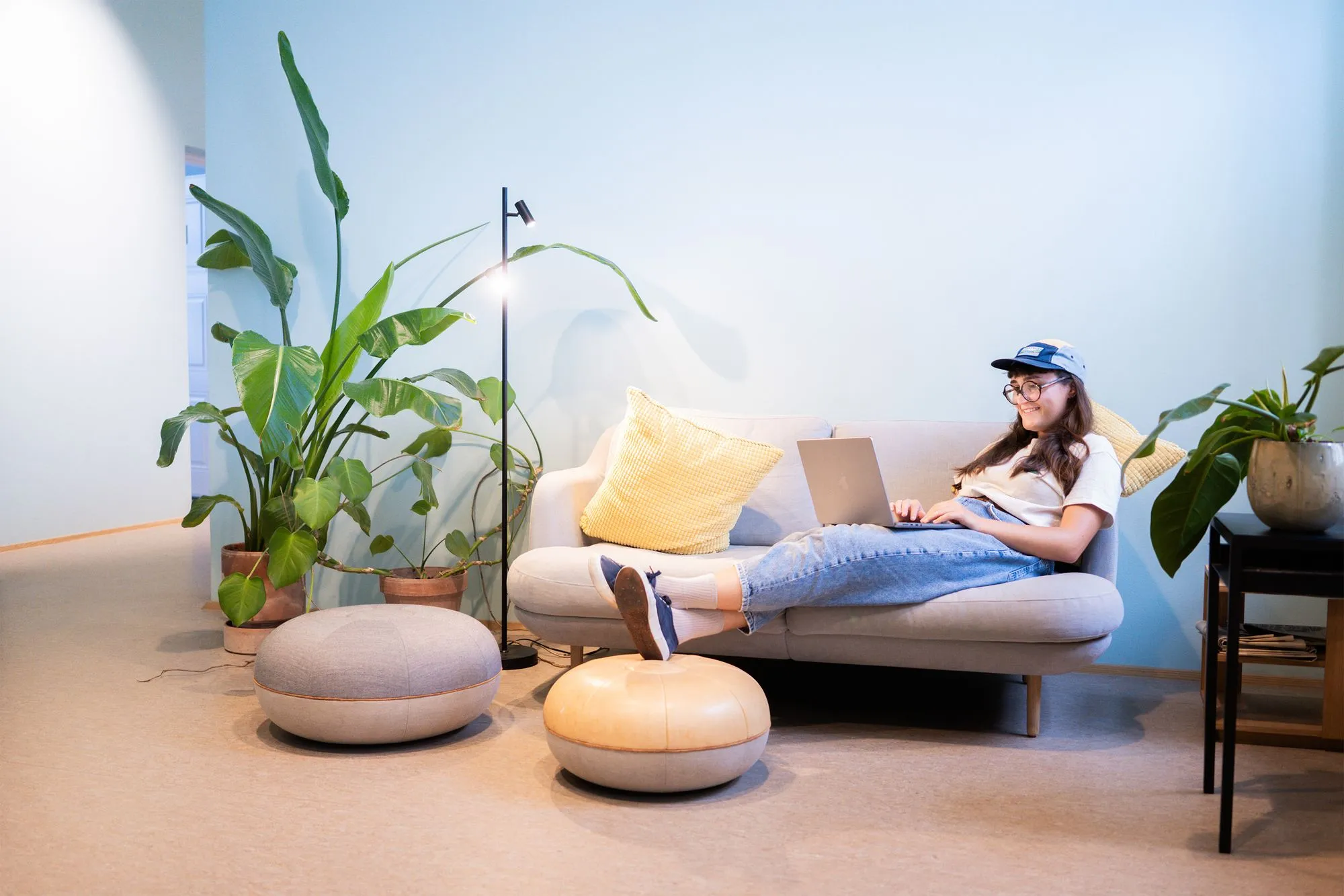 Person relaxing on a couch with a laptop in a cozy, plant-filled space.