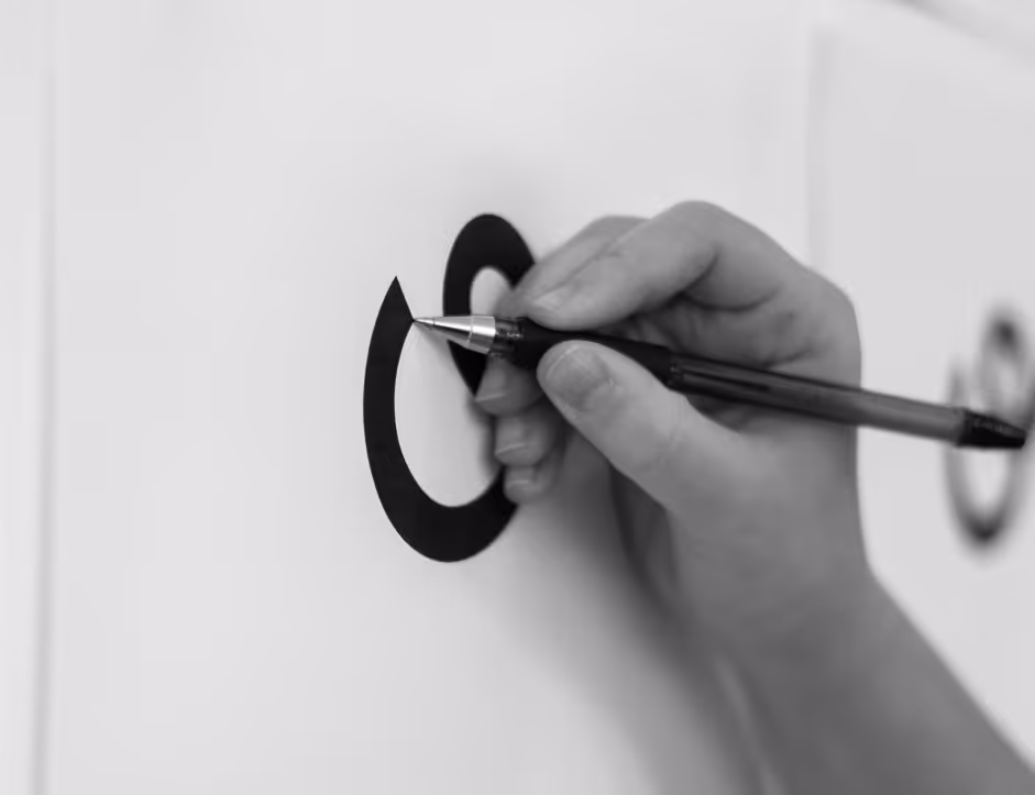 Hand holding a pen and drawing the logo sketch on a white surface.