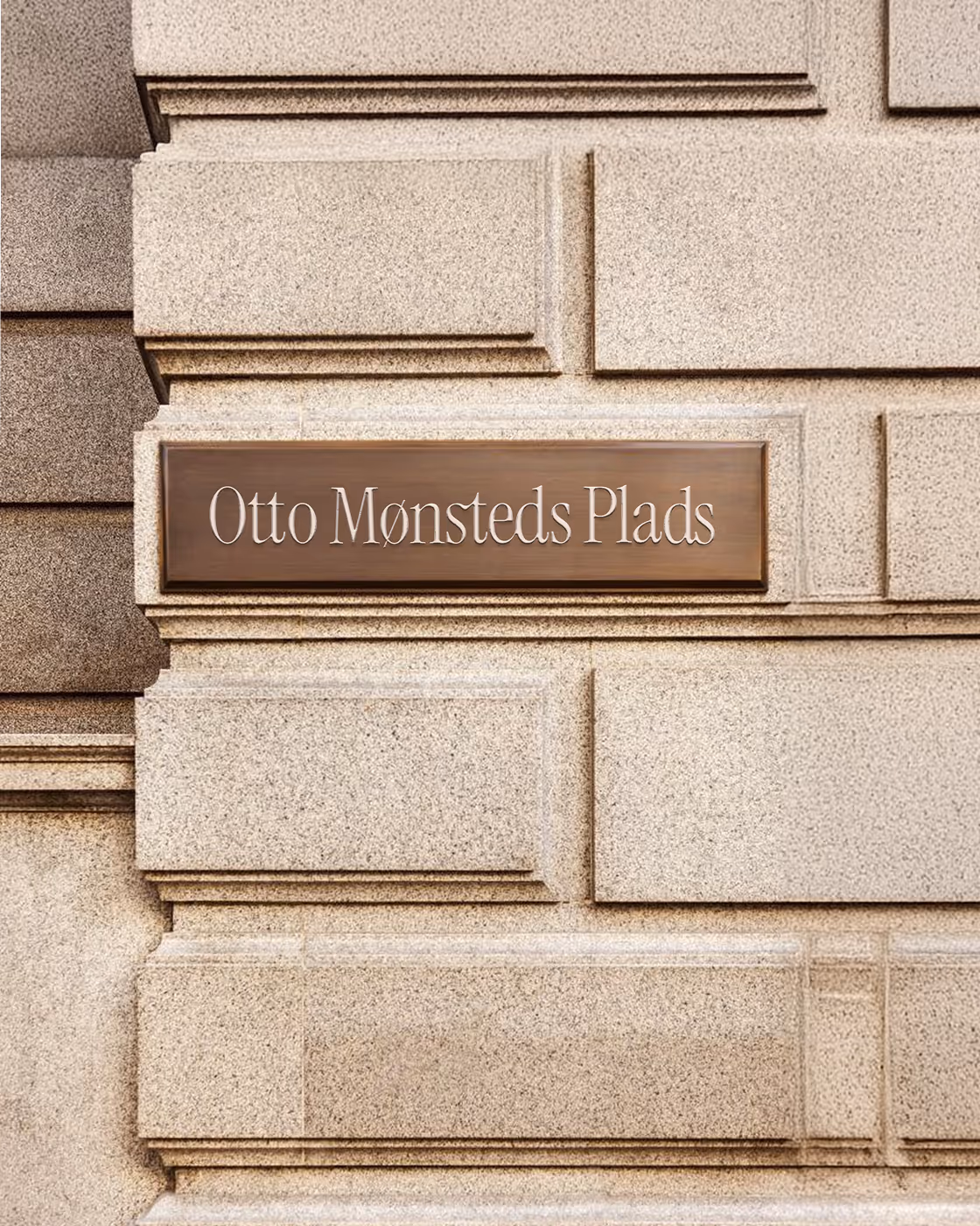 Brown street sign reading 'Otto Mønsteds Plads' mounted on a beige stone wall with rectangular carvings.