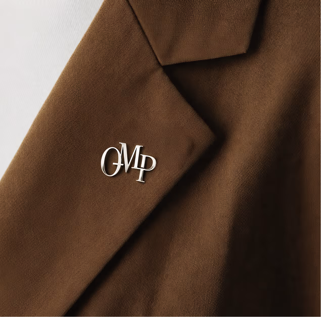 Silver pin with intertwined letters 'OMP' attached to the lapel of a brown blazer.