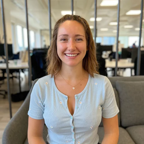 Smiling woman with brown hair wearing light blue buttoned shirt sitting on a gray couch in an office.