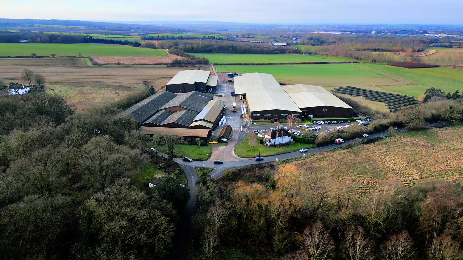 An aerial view of large industrial buildings surrounded by fields and trees on a cloudy day.