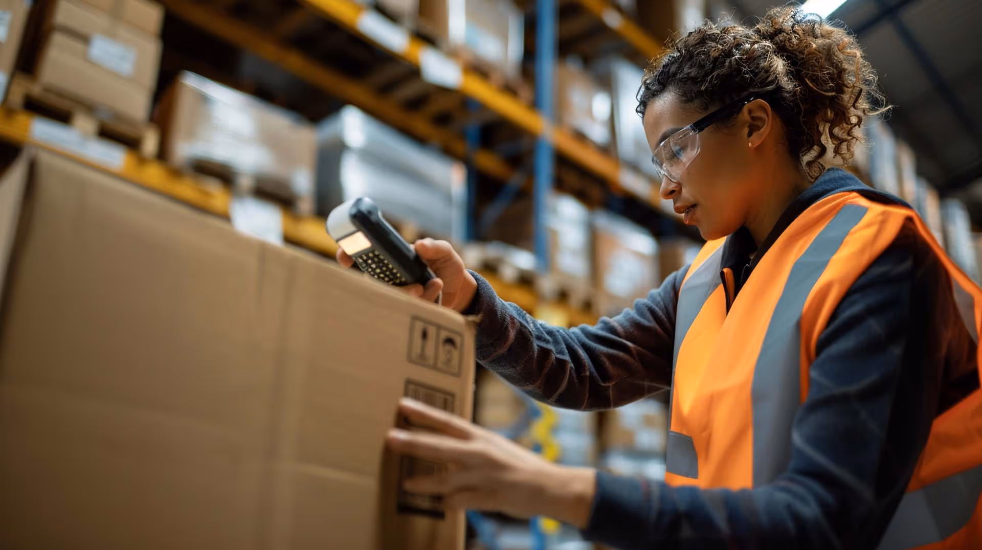 A warehouse worker in an orange vest scans a large cardboard box with a handheld device.