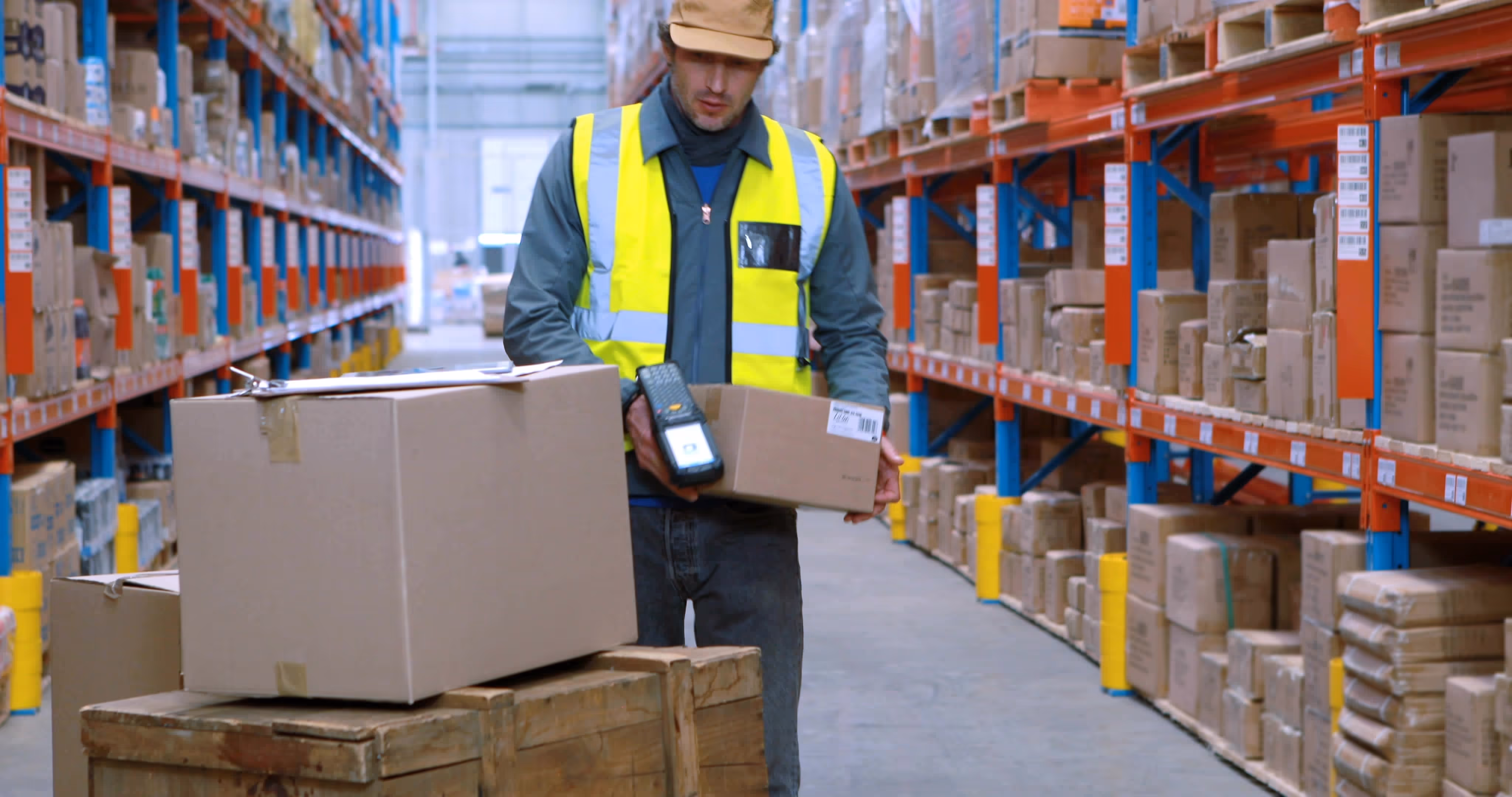 A worker in a high-visibility vest lifts boxes in a warehouse aisle filled with shelves.