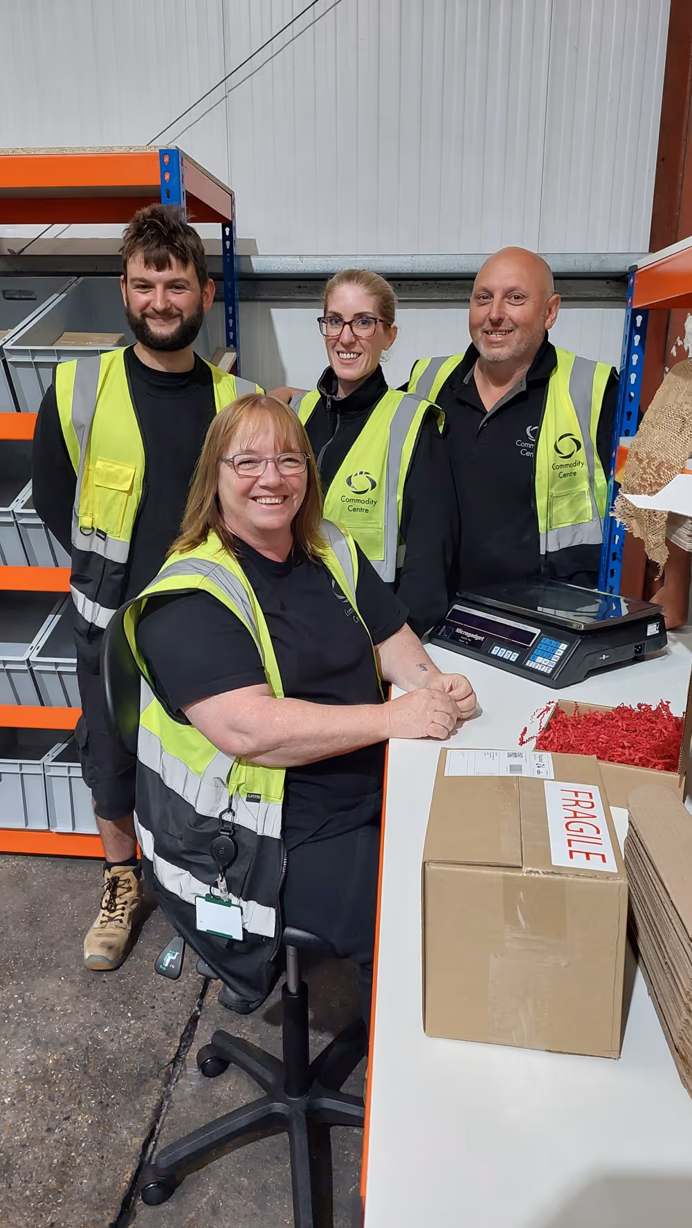 Four people in safety vests are smiling in a warehouse, standing by boxes labeled "fragile."