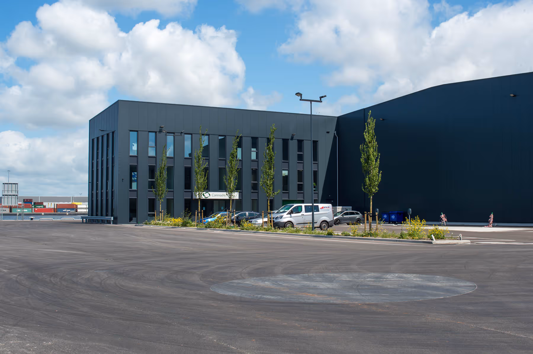 Un immeuble de bureaux gris moderne avec plusieurs voitures en stationnement et de jeunes arbres à proximité.