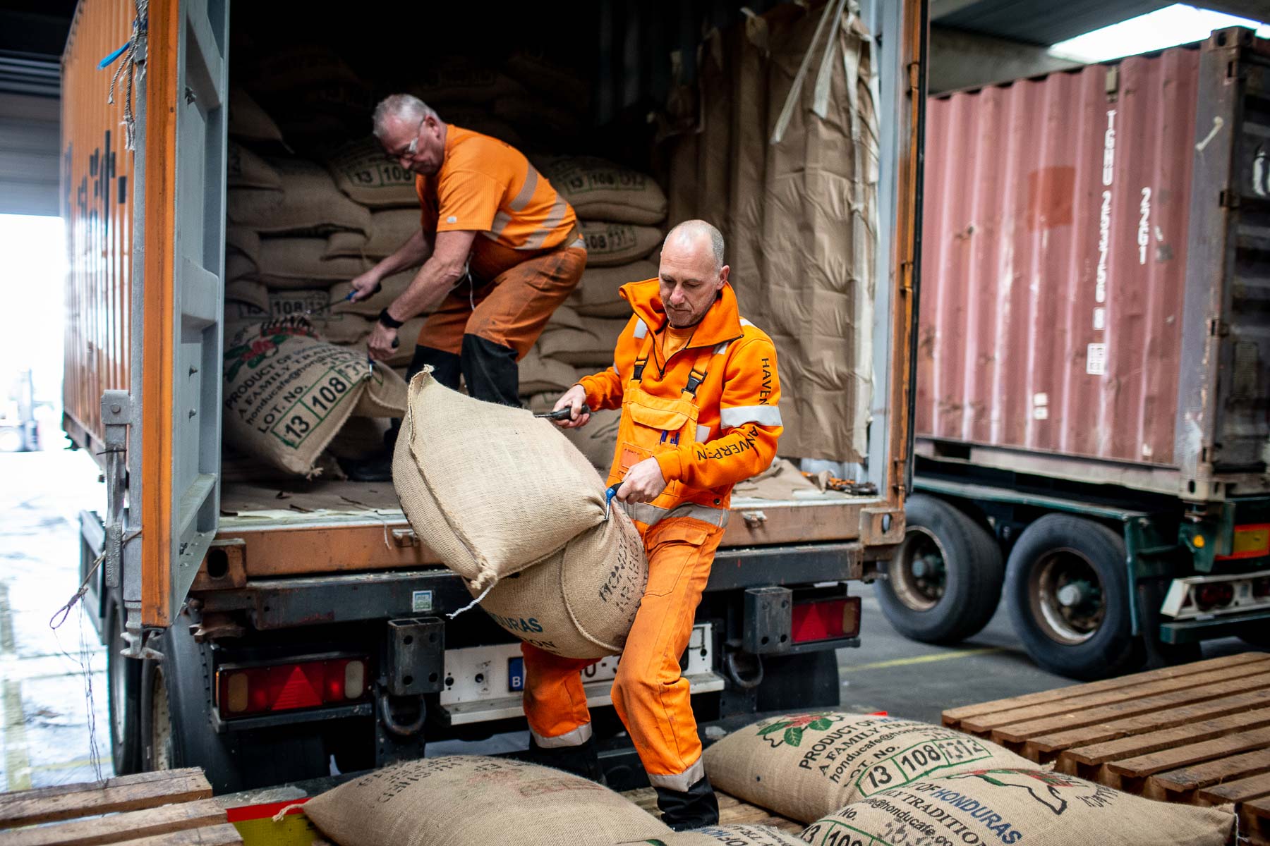 2 men in orange overalls unloading bags from the back of a truck 
