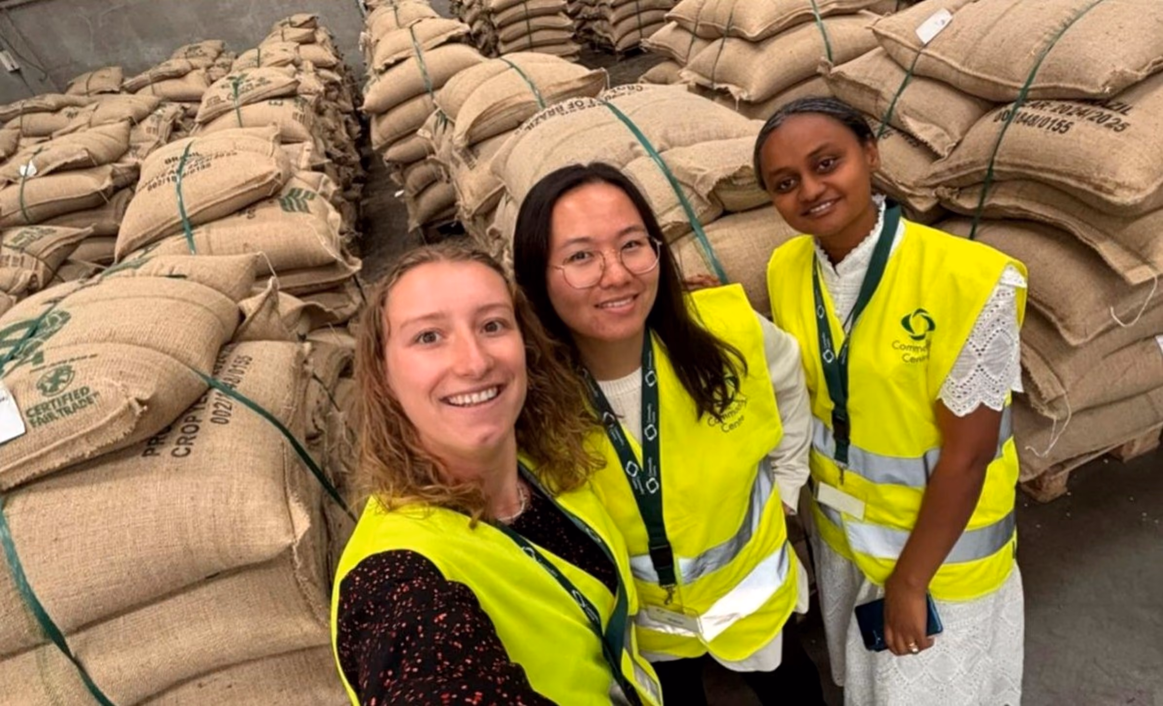 Three people wearing high-visibility Commodity Centre safety vests smile for a selfie inside a warehouse filled with neatly stacked hessian sacks of agricultural commodities. The sacks are arranged in large piles on either side, creating aisles behind the group, highlighting the scale of the storage facility.