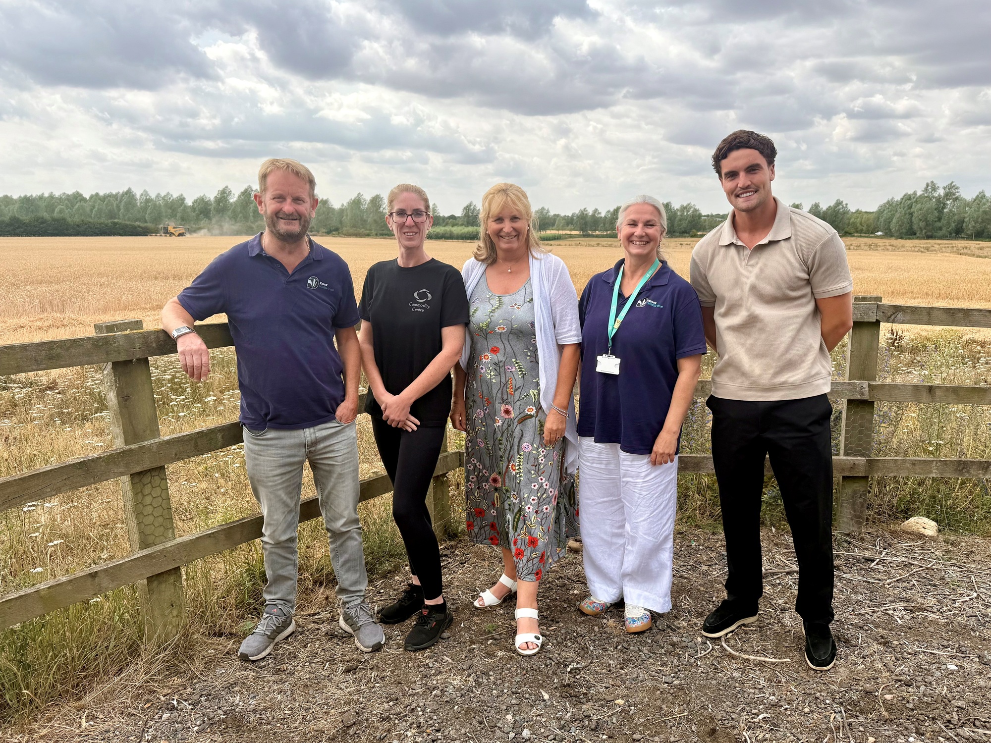 Three people stand beside a wooden fence in a rural field with a large solar farm in the background under a blue sky with scattered clouds. One person holds several brochures while the group smiles at the camera, highlighting a partnership between environmental conservation and renewable energy.