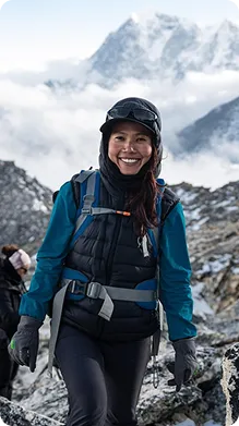 Naomi; A Smiling woman wearing outdoor hiking gear, standing on rocky terrain with snowy mountains and clouds in the background.