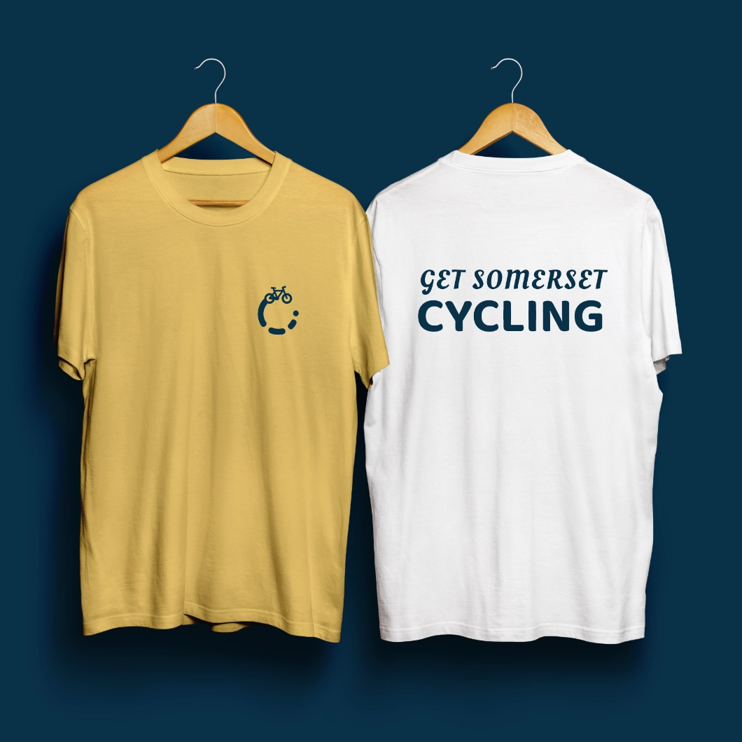 'Get Somerset Cycling' logo and word mark. The logo features a minimal bicycle with a progressing circle around it. Printed on a t-shirt.