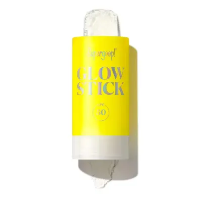 Supergoop Glow Stick SPF 50 chemical sunscreen stick with dewy highlighter finish in yellow packaging on sandy background with sunglasses and palm leaves