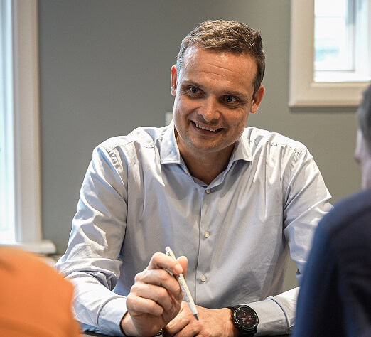 Mand i lyseblå skjorte smilende og peger med en pen under et møde i et lyst kontormiljø.