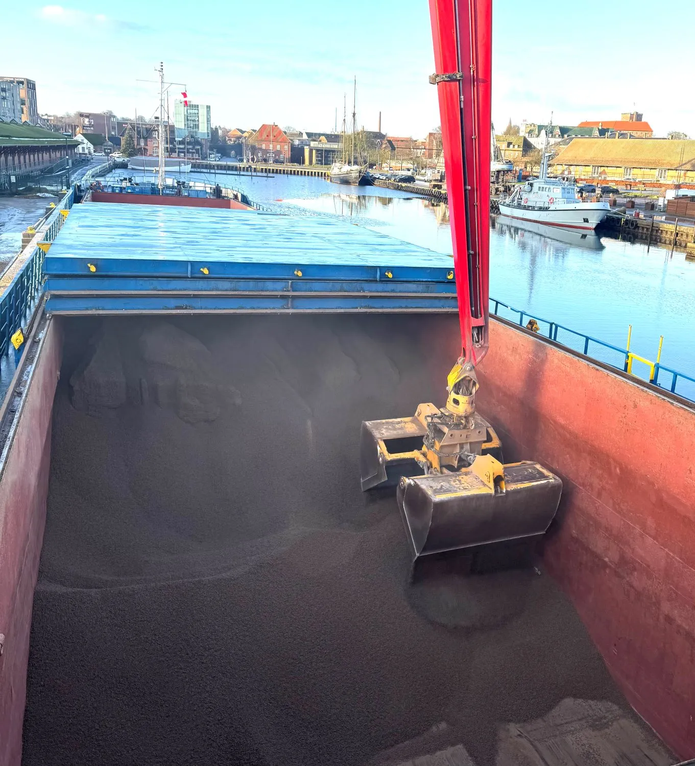 Stor kran med gribeklo læsser bulk materiale fra et skib i en havn med bygninger og både i baggrunden.