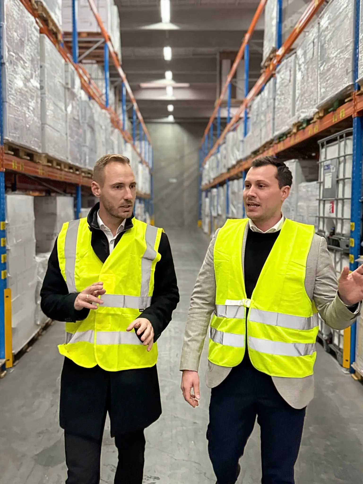 Two men wearing yellow vests walk into a warehouse.