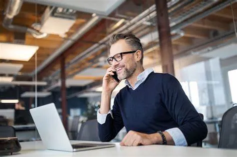 Business man in glasses and navy sweater smiling while talking on phone at desk with laptop in modern office.