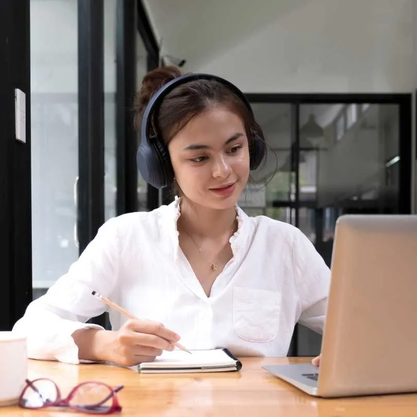 Business woman wearing headphones writing in a notebook while looking at a laptop in a modern office.