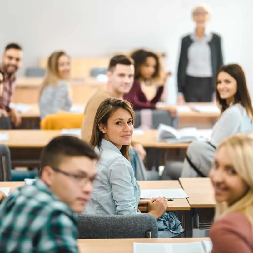 Attendees sitting at desks in a classroom, some looking toward the camera, with a teacher standing in the background.