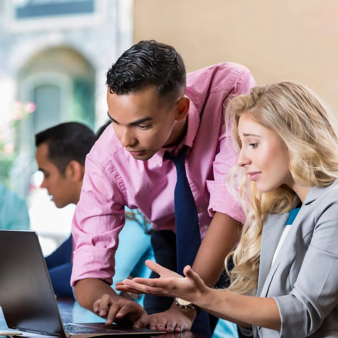 Business man in pink shirt and blue tie assisting a blonde woman in a gray blazer with a laptop, while another person works in the background.