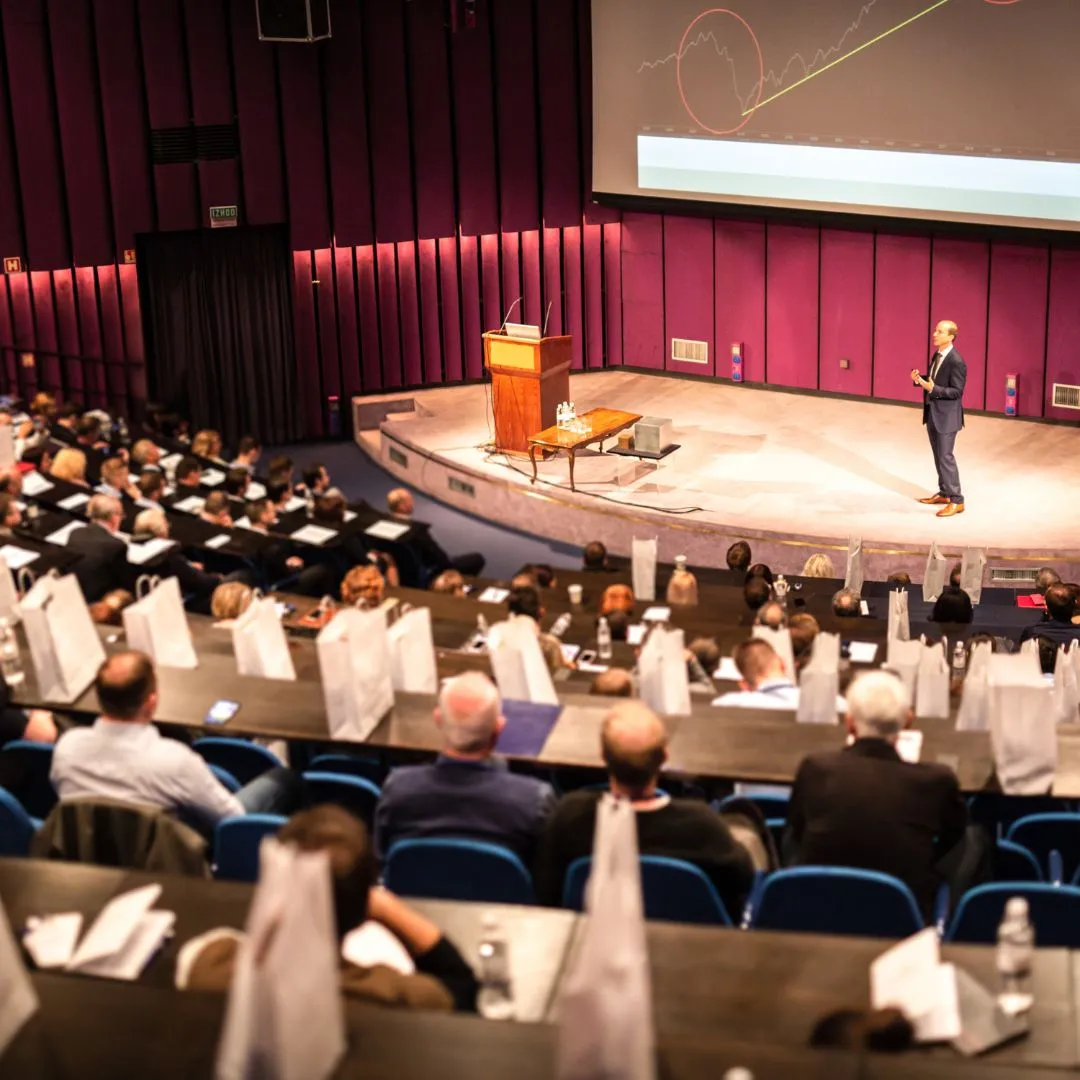 Audience seated in an town hall listening to a speaker in a suit presenting a graph on a large screen.