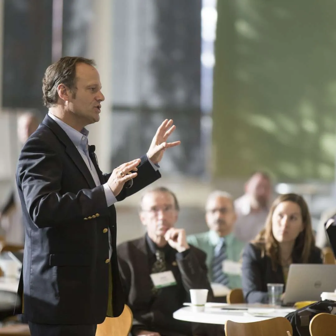 Business man in a dark blazer speaking and gesturing to an audience seated at tables in a conference or seminar setting.