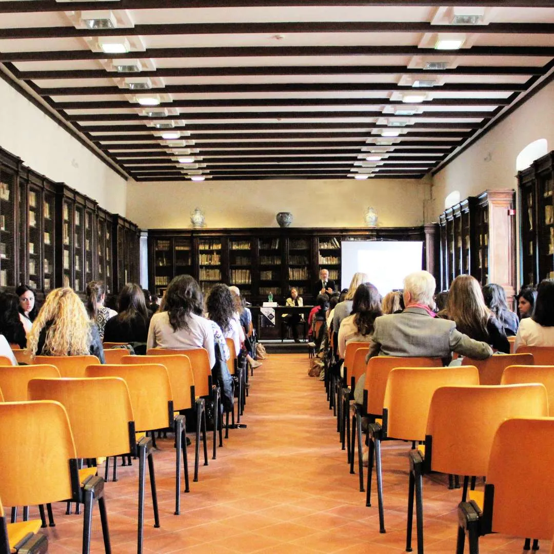 Audience seated in rows of wooden chairs facing a speaker presenting in a library room with bookshelves and ceiling beams.