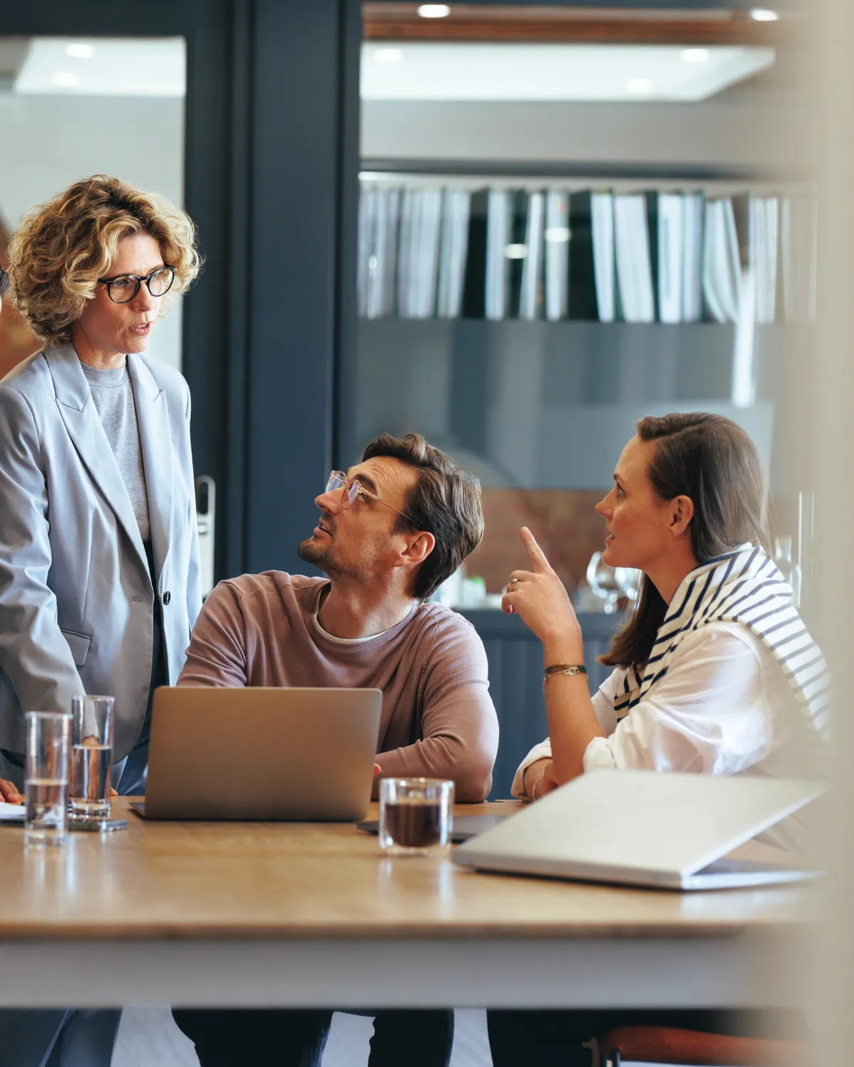 A man and two women having a discussion in an office with laptops and glasses on the table.
