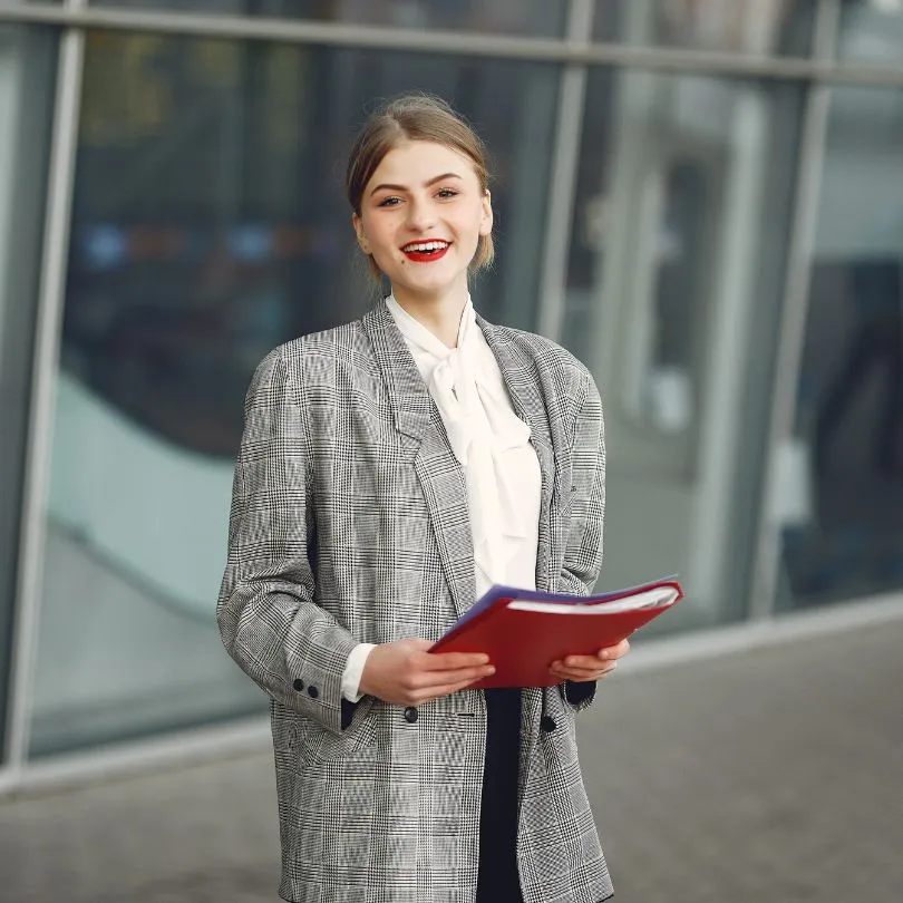 Smiling young professional woman wearing a gray checkered blazer and white blouse holding a red folder standing outdoors.
