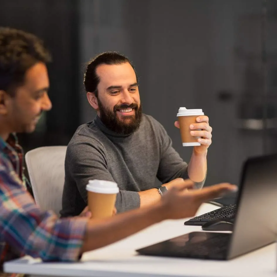Two business men sitting at a desk with coffee cups, smiling and looking at a laptop screen.